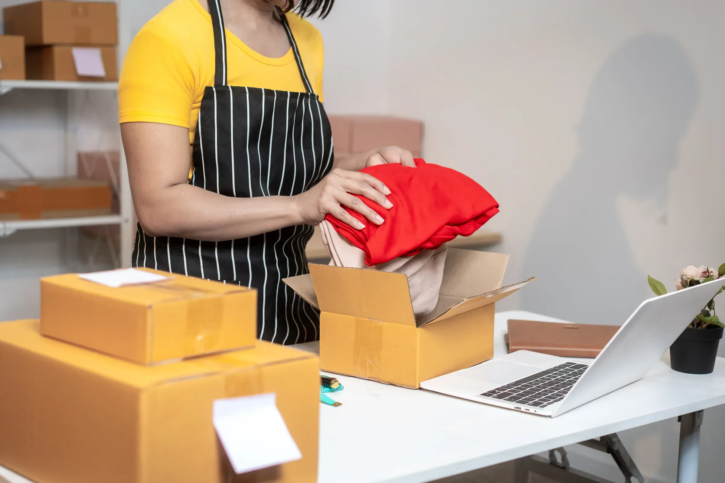 Woman packing folded clothes into a cardboard box at a home workspace for online order shipping, with laptop,, amazon sellers going dtc ecommerce