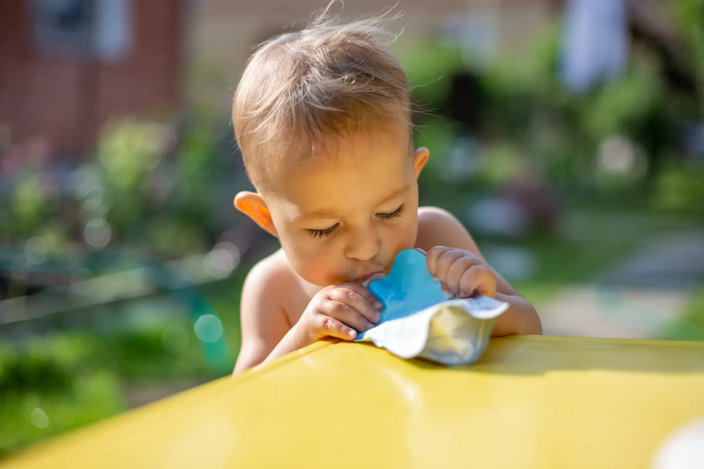Baby eating fruit puree in pouch and looking into the food in front of the yellow table. on the background is, baby & toddler food ecommerce
