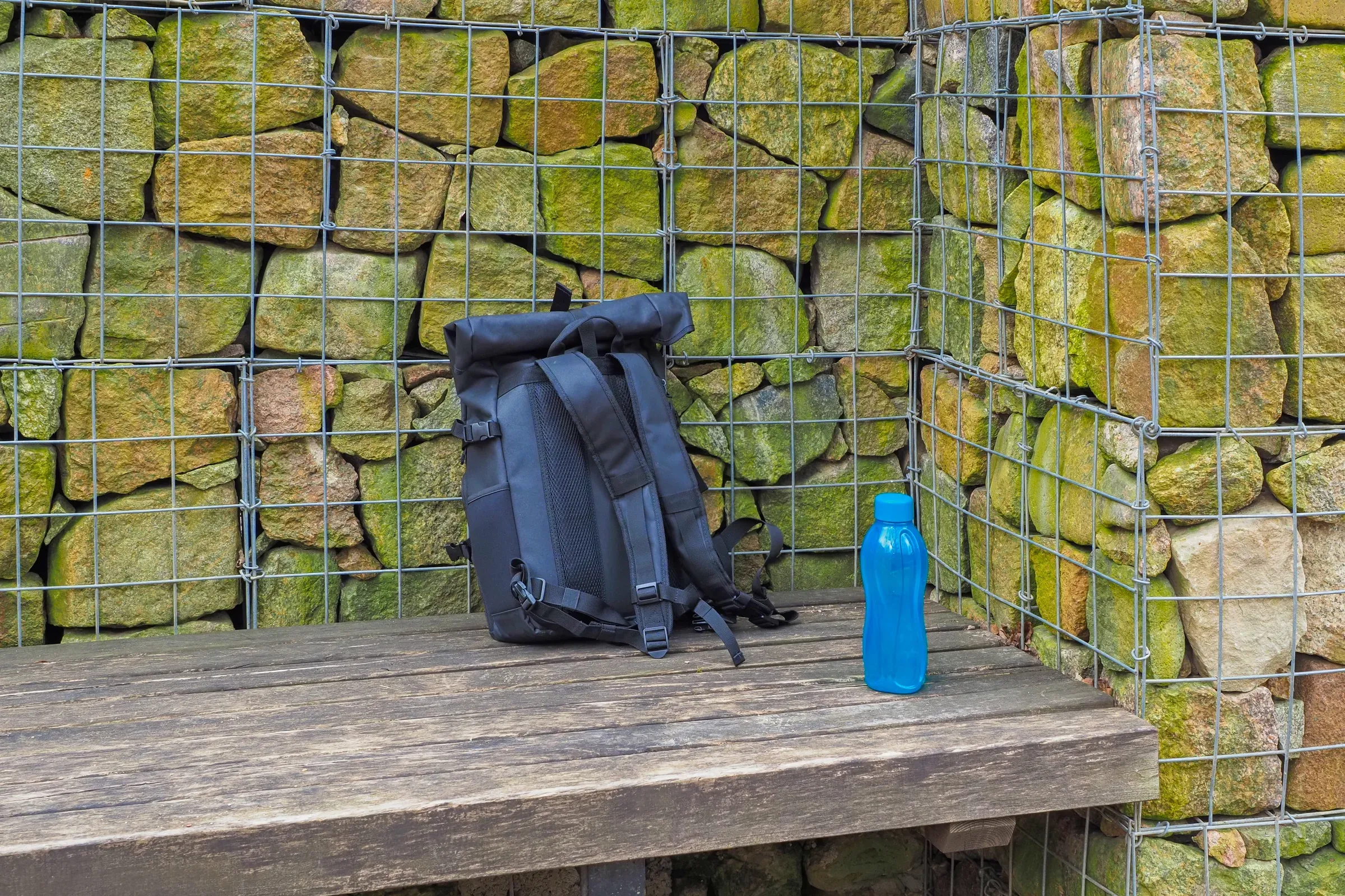 Black rolltop backpack and blue reusable water bottle on a wooden bench against a gabion stone wall, backpacks ecommerce