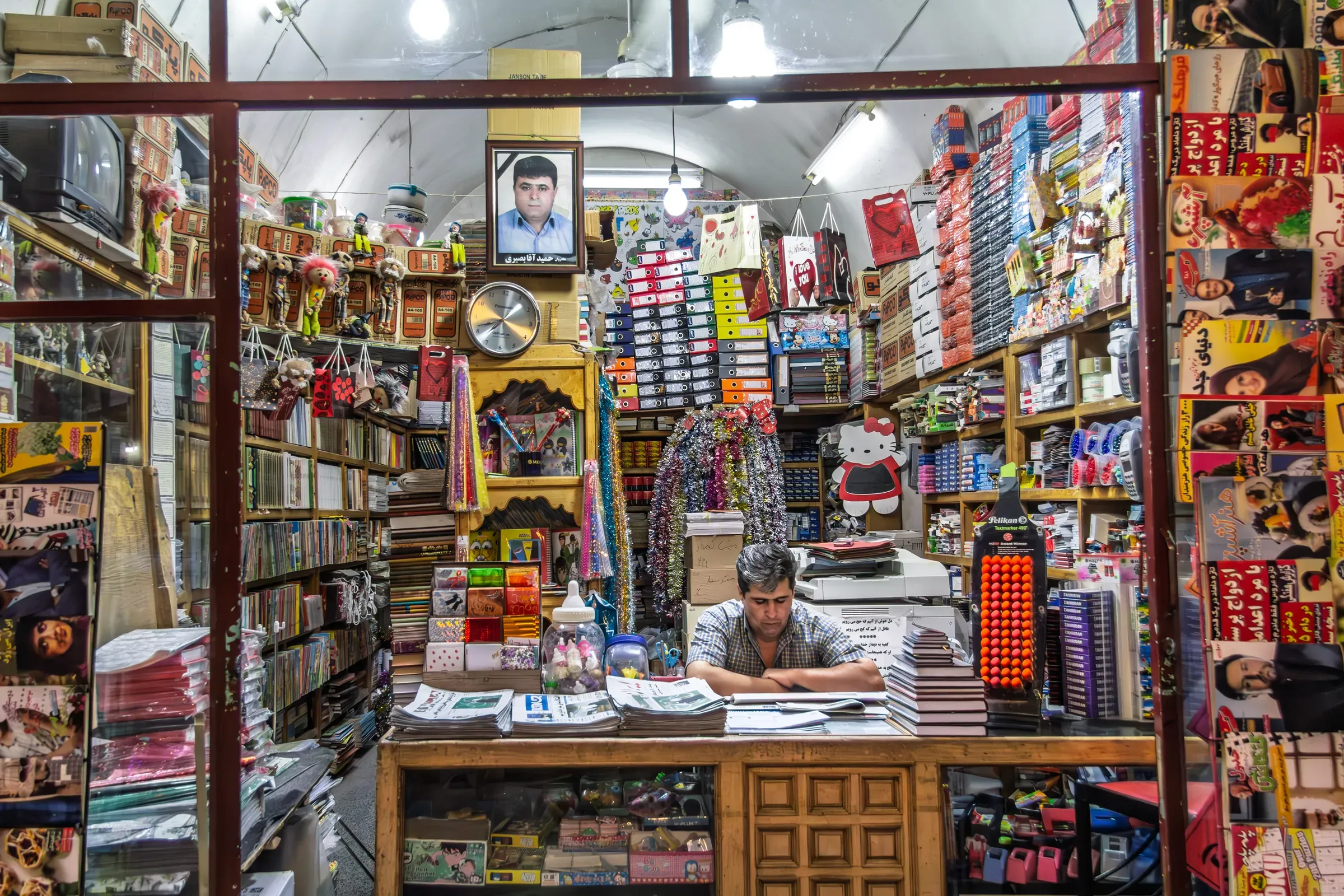 Shopkeeper sits behind a counter in a packed school and office supplies store in Yazd, Iran, through the, books, stationery & paper ecommerce