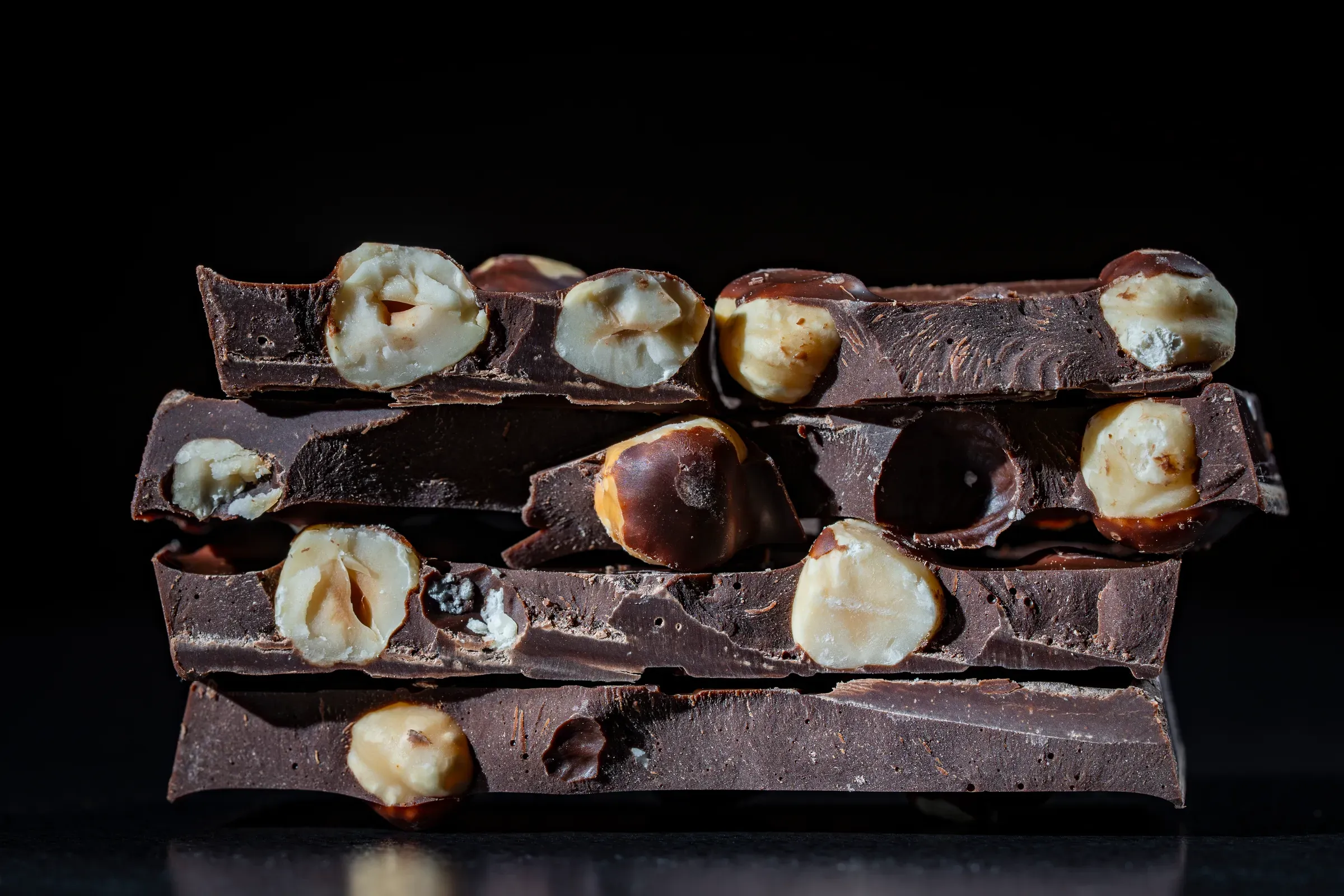 Stack of dark chocolate bars with whole hazelnuts on black background. Closeup of broken dark chocolate, chocolate & confectionery ecommerce