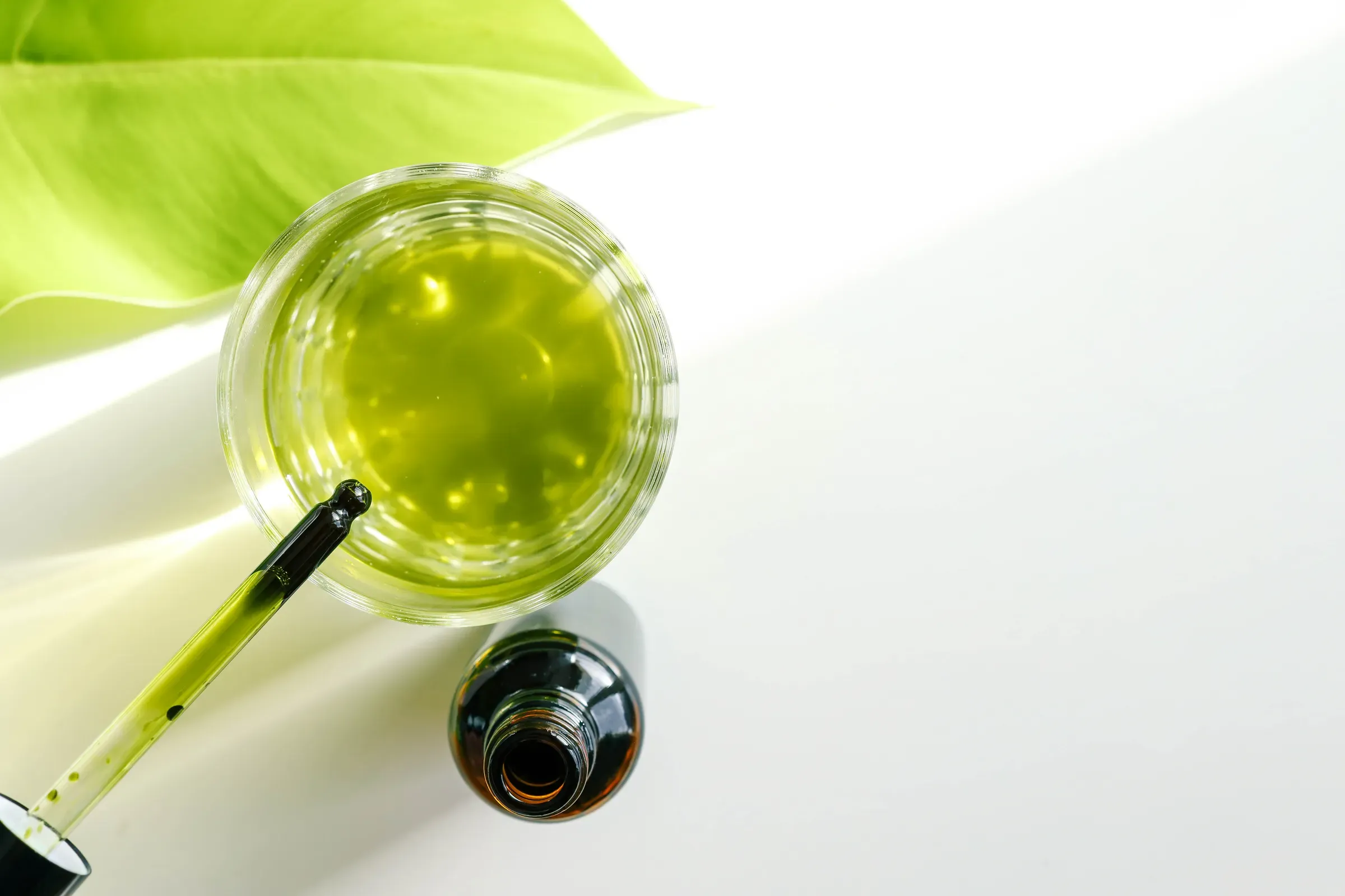 Minimalistic composition with a green glass dropper, natural oil in a transparent dish, and a large green, clean skincare ecommerce