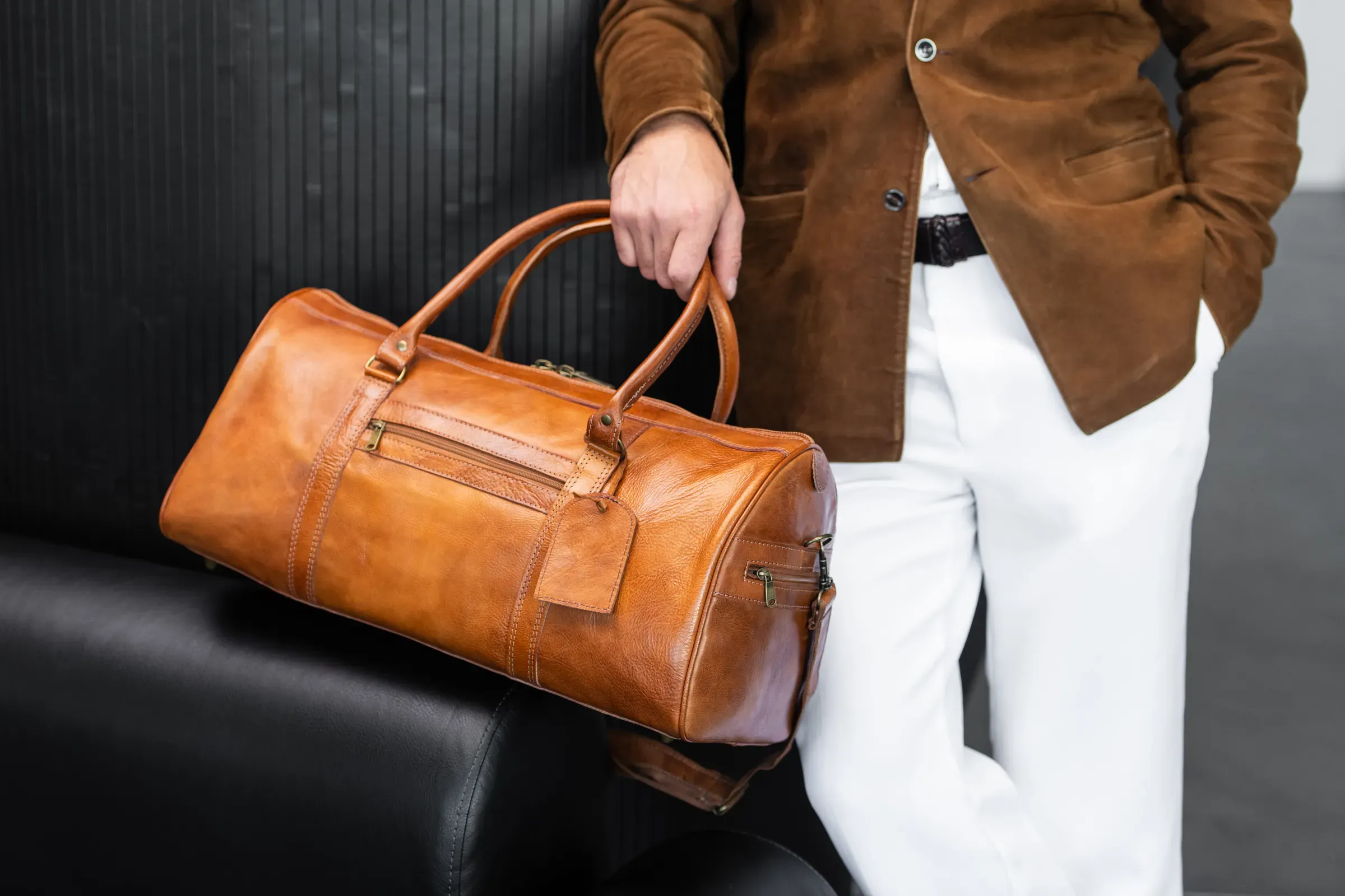 Close up of a man holding a premium tan leather duffel bag wearing a suede blazer and white trousers, duffel & weekender bags ecommerce