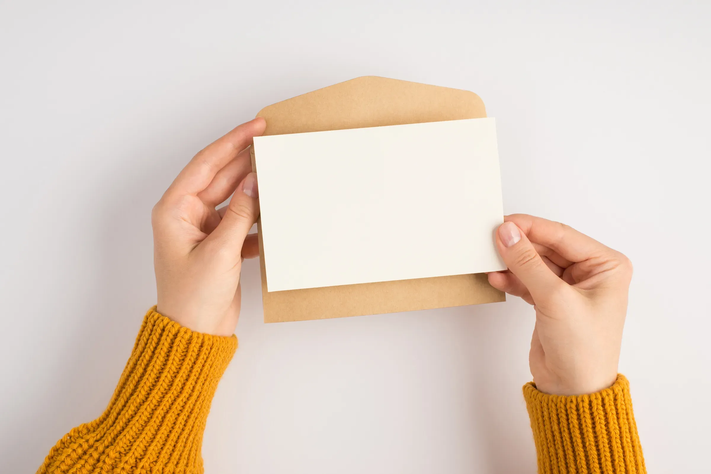 First person top view photo of female hands in yellow sweater holding open craft paper envelope and white, greeting cards ecommerce