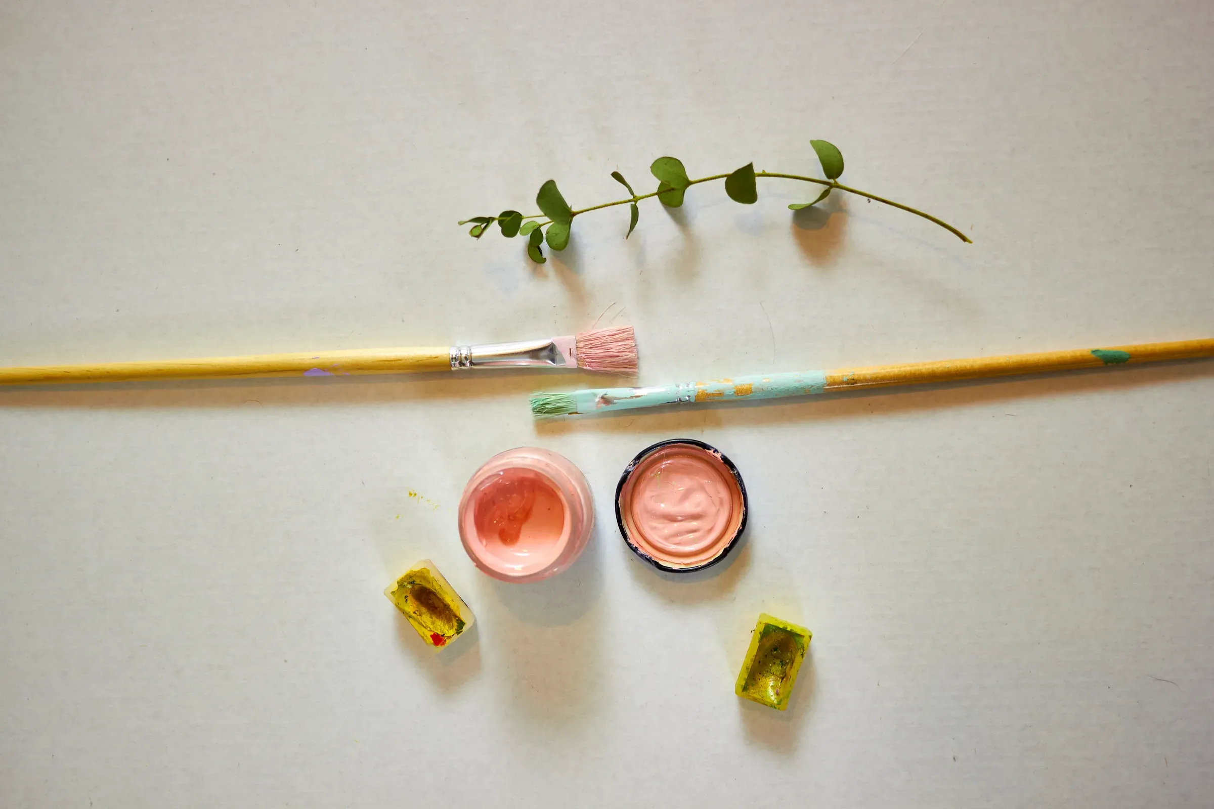 Creative flat lay of watercolor paints, brushes, and green branch on light background. Artistic workspace, hobby, craft & creative ecommerce