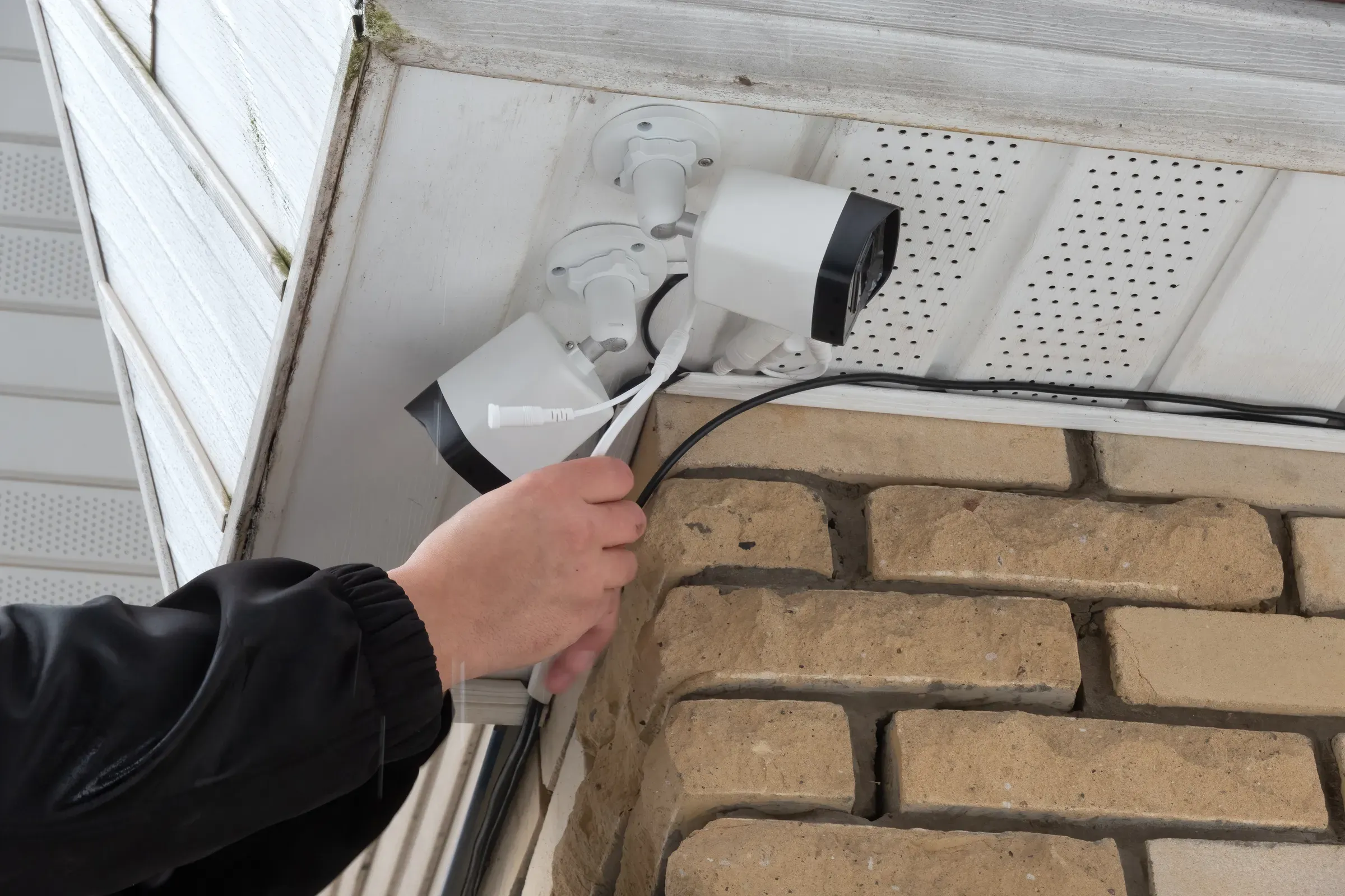 A person's hand connects wires to new security cameras mounted under white building eaves, next to a brick, home security cameras ecommerce