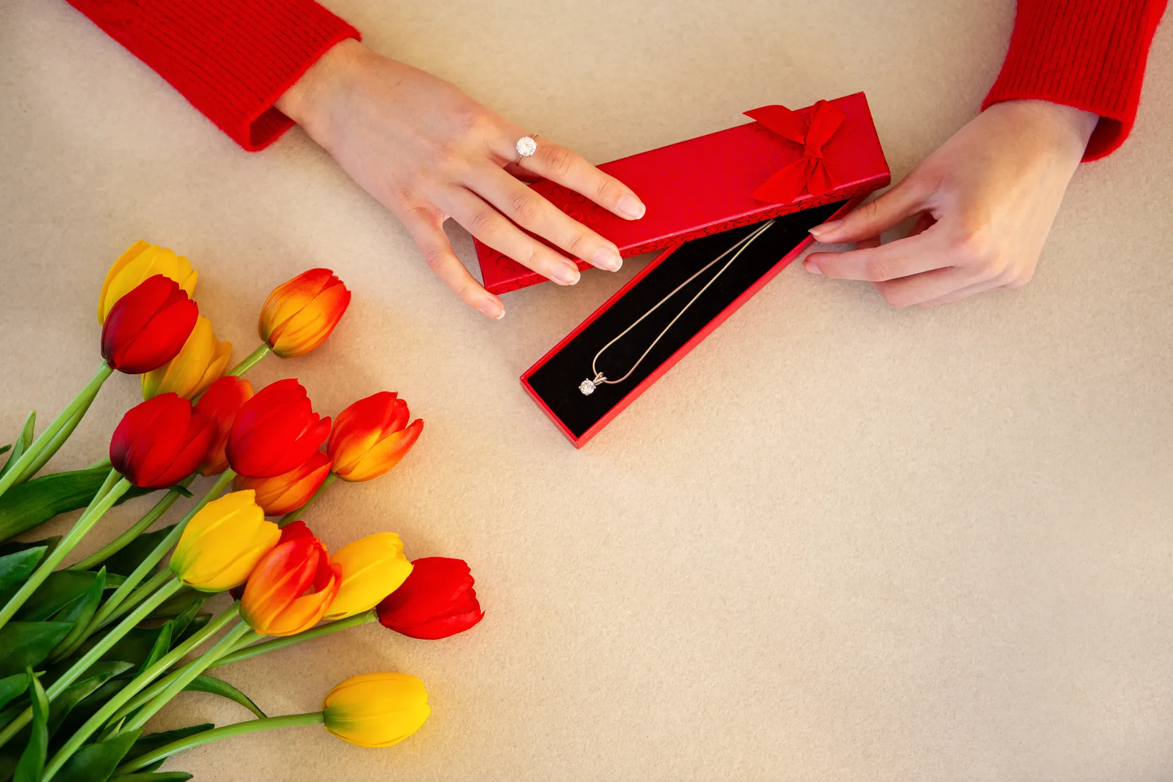Opening jewelry gift box with tulips. Top view of hands opening a red jewelry box with a necklace beside, jewelry subscription ecommerce