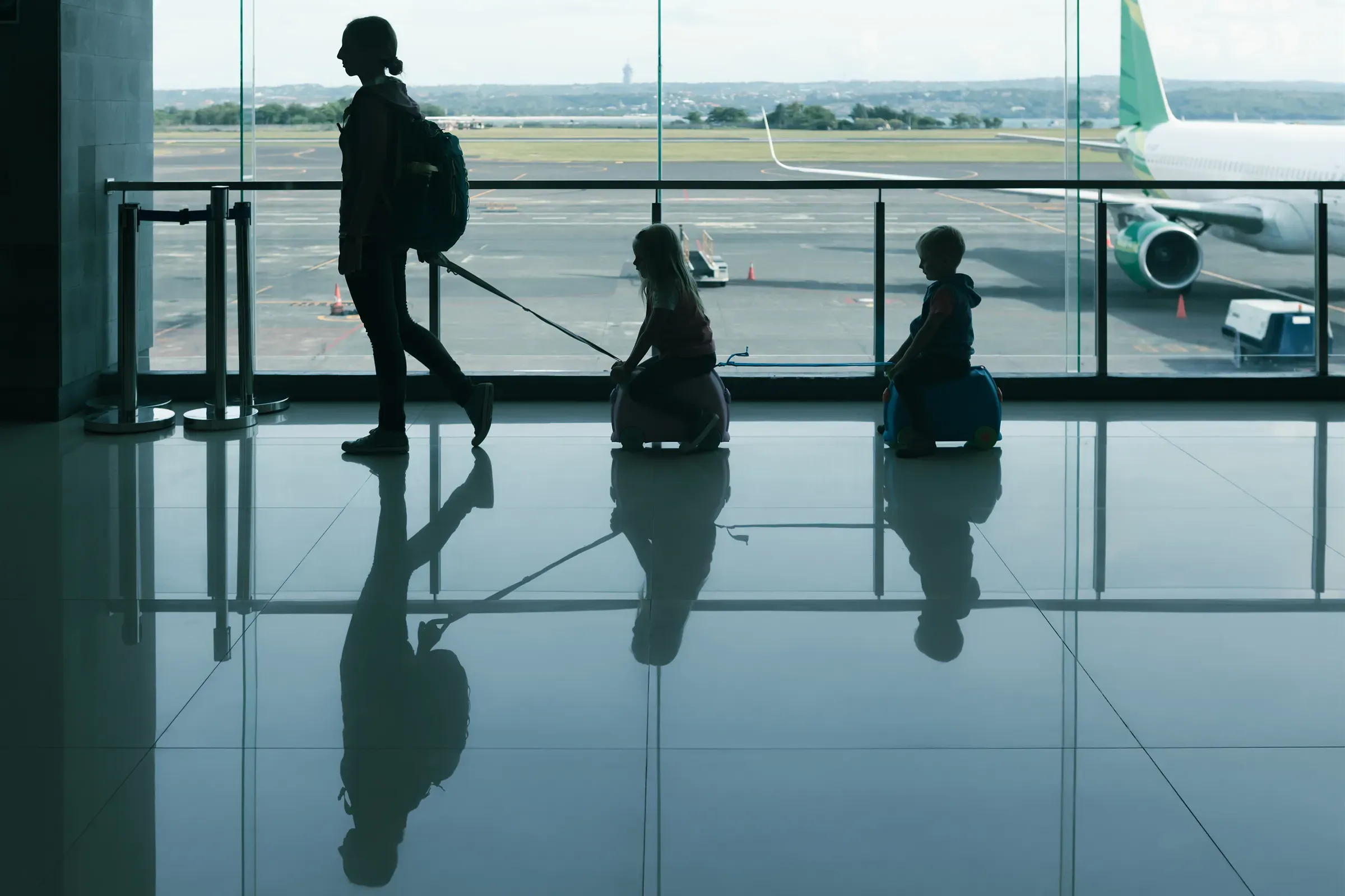 A mother pulls two children on small ride-on luggage through airport terminal, silhouetted against large, kids luggage & travel ecommerce
