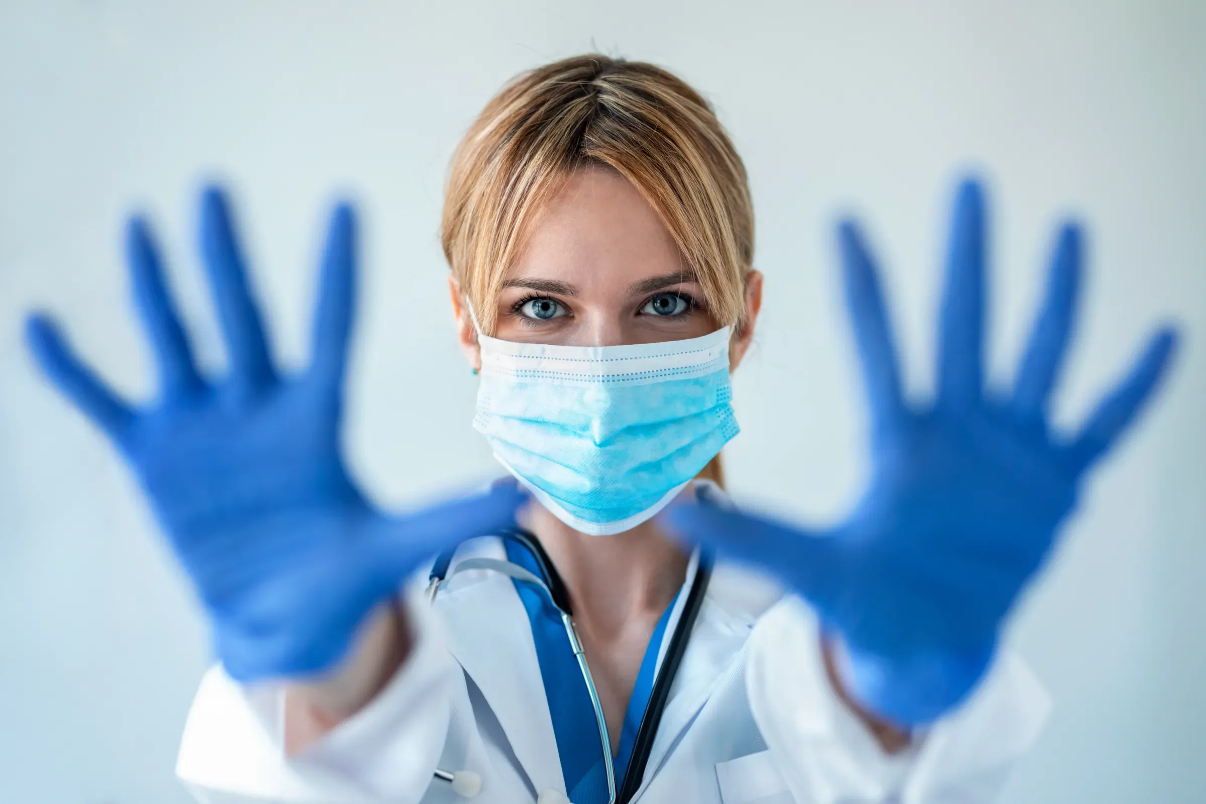 Female doctor with a medical mask showing hands to camera while wearing a blue nitrile gloves over white, medical & dental supply ecommerce