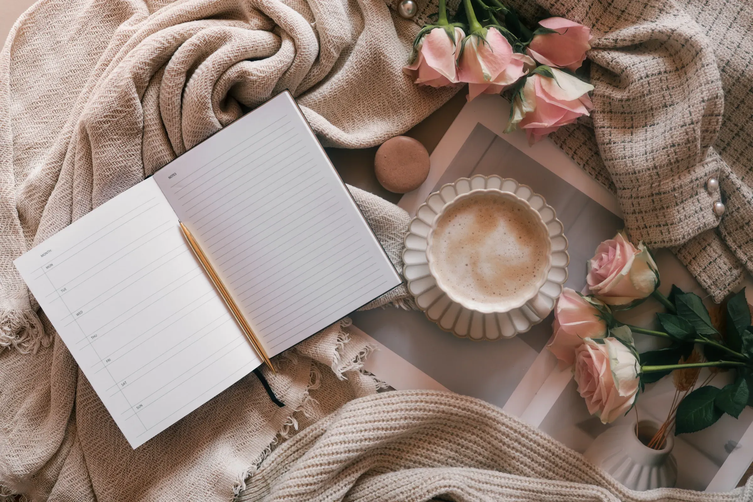Flat lay of an open weekly planner with a pen, coffee cup, pink roses, and cozy neutral fabrics, planners & journals ecommerce