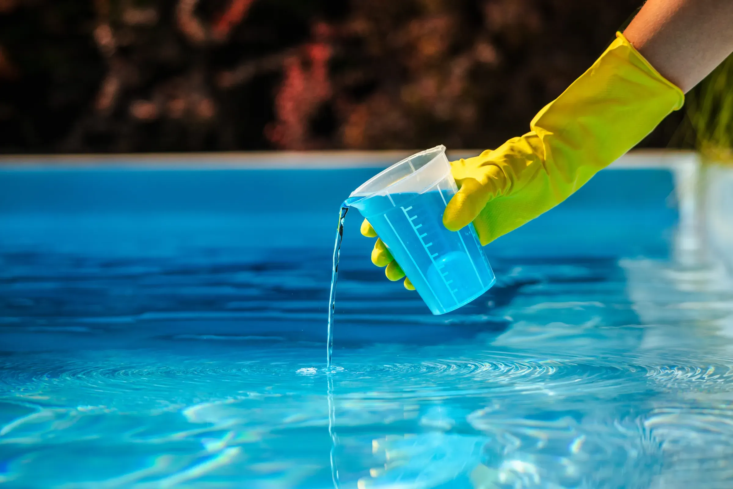 Hand with yellow protective glove pouring blue chemical liquid from measuring cup into swimming pool water, pools & pool supplies ecommerce