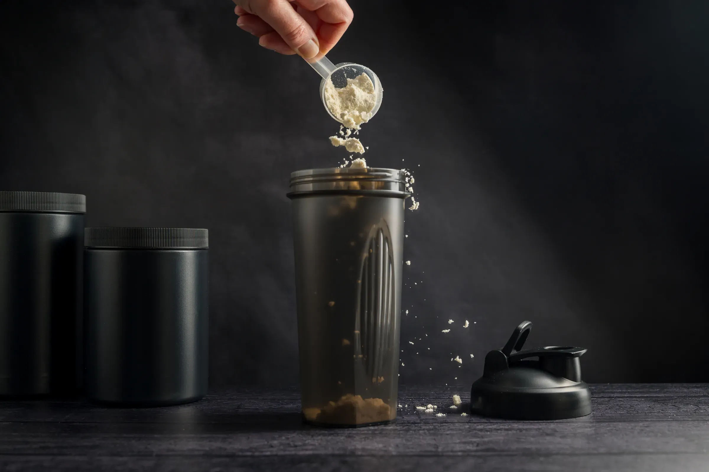 Protein drink. Hand pouring protein powder in a black shaker. Plastic tube on dark background. Body building,, sports nutrition & protein ecommerce