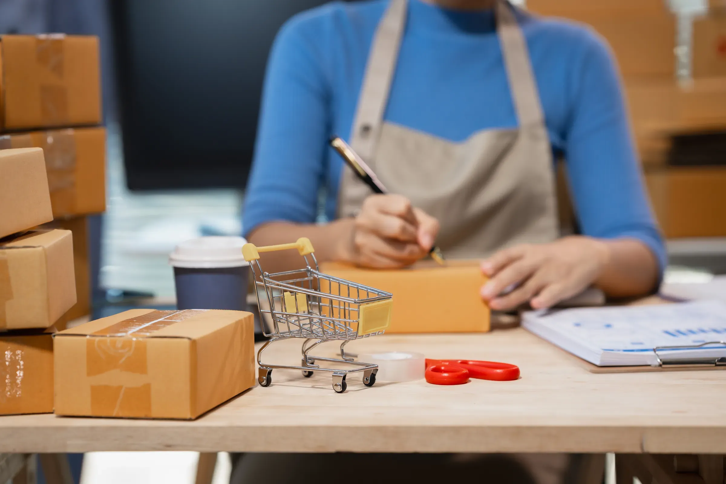 Small business owner packing orders at a desk with cardboard boxes, mini shopping cart, coffee cup and, store types & business models ecommerce