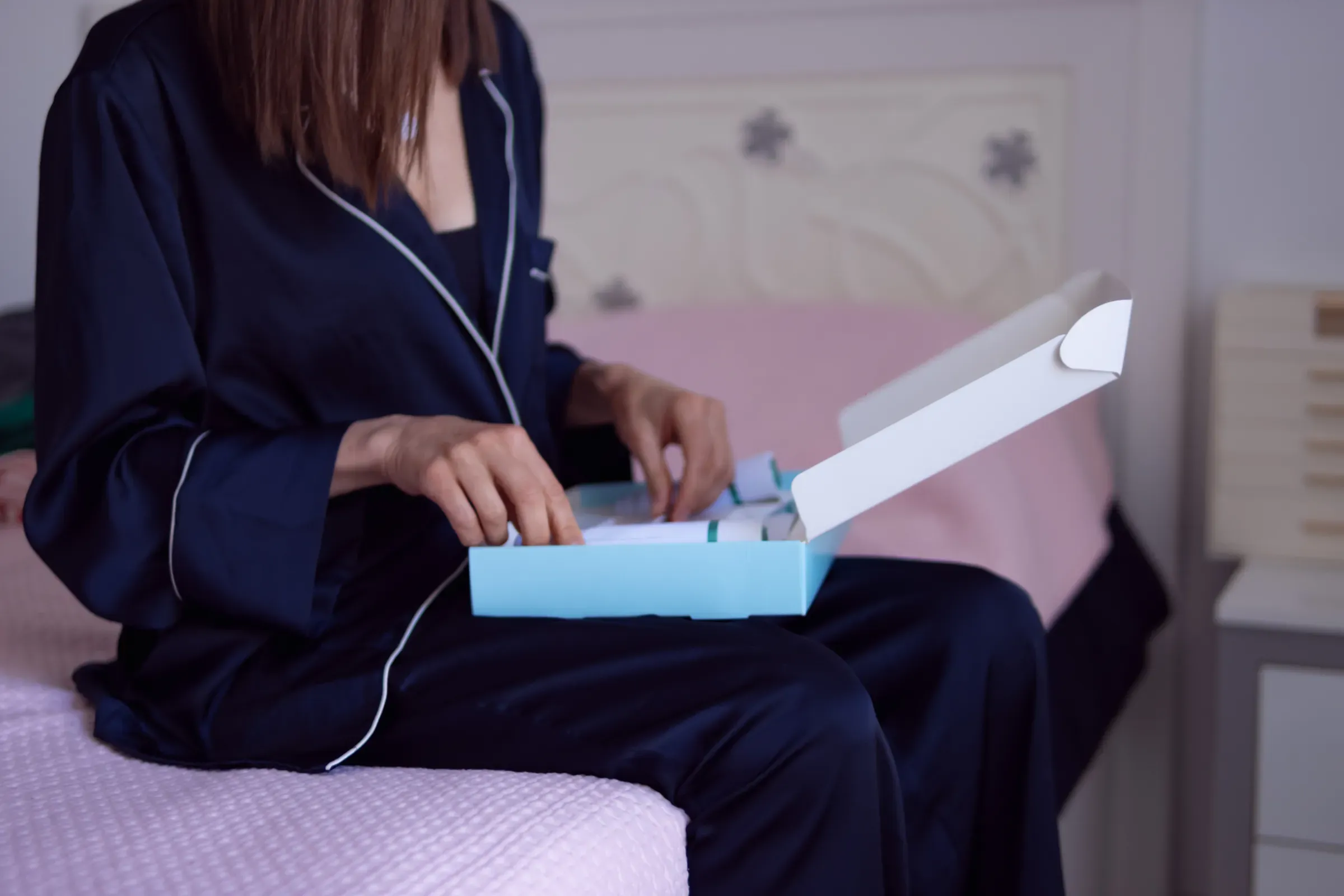 Woman in pajamas unboxing a blue gift box on her bed, discovering a surprise package as part of a relaxing, subscription gift boxes ecommerce