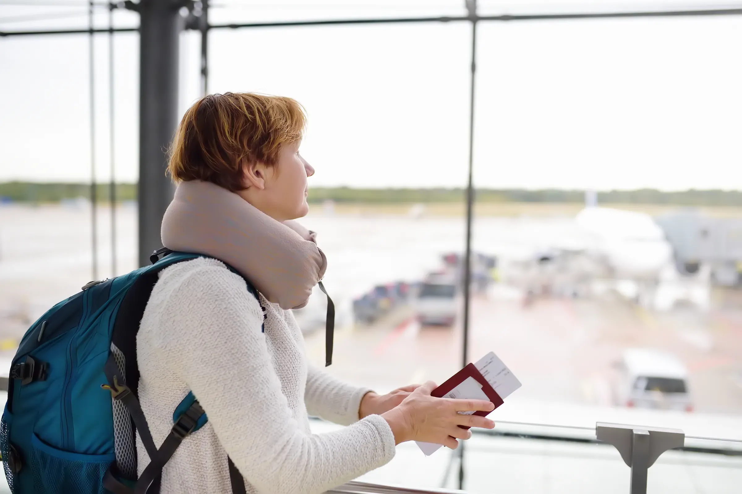 Woman with a backpack and a travel pillow looks on the plane in window on waiting area of the airport, travel pillows & comfort ecommerce
