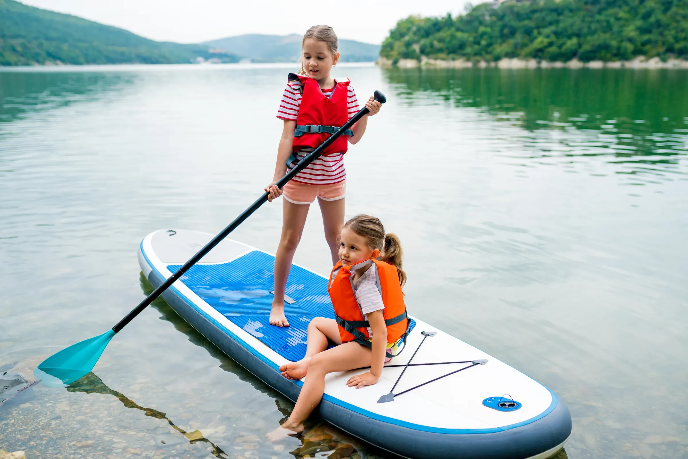 Girls padding on stand up paddle boarding on lake district. Children in swim life vest learning, water sports ecommerce