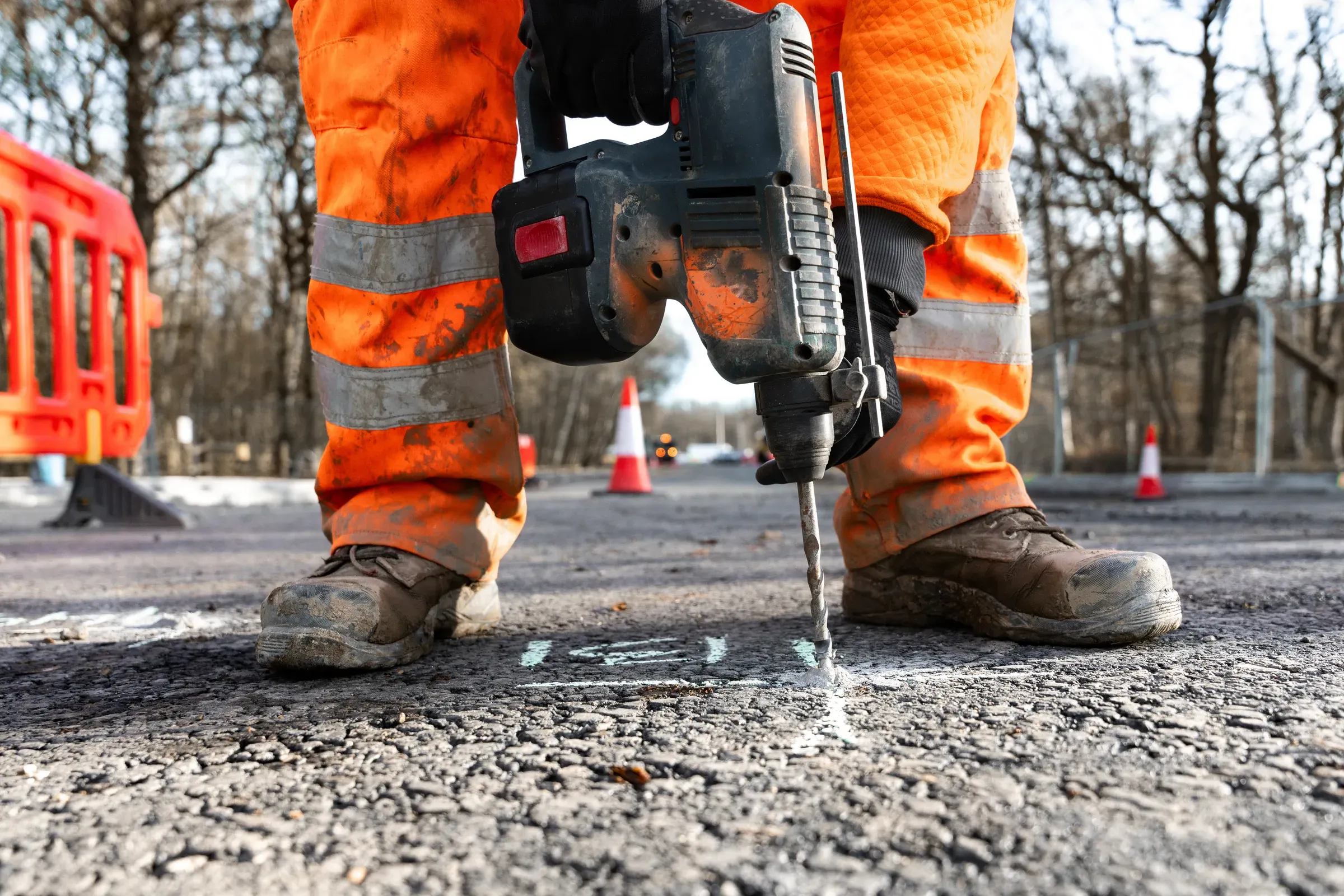 Construction worker in orange clothes and safety boots using cordless drill to break asphalt on road project, work boots ecommerce